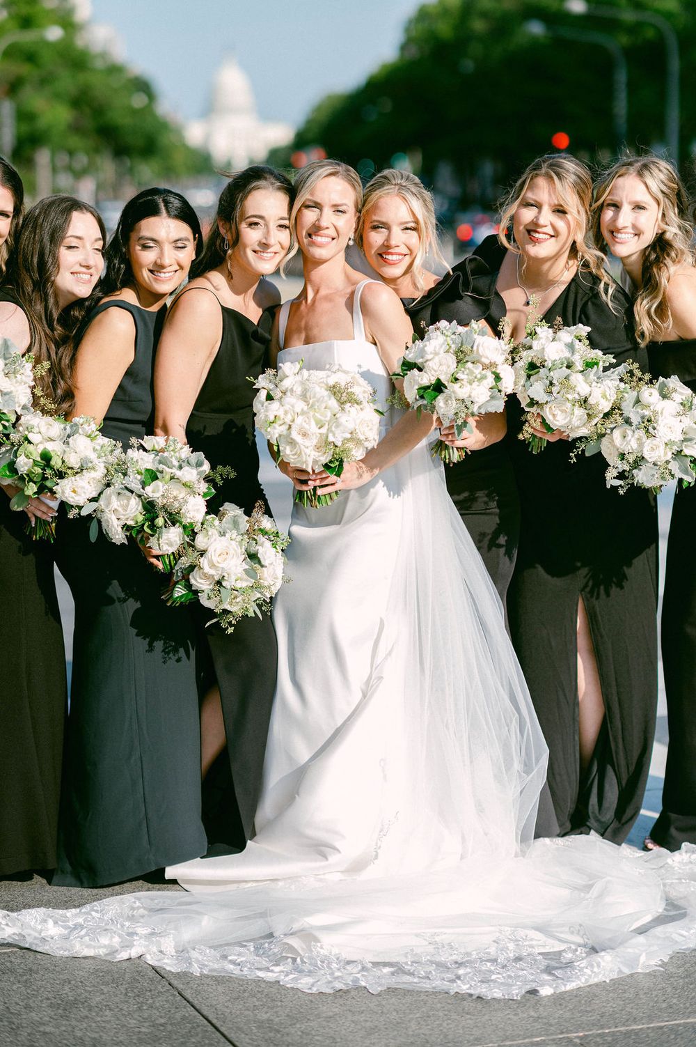 Wedding party in black bridesmaid dresses and white wedding gown pose together holding elegant white floral bouquets.