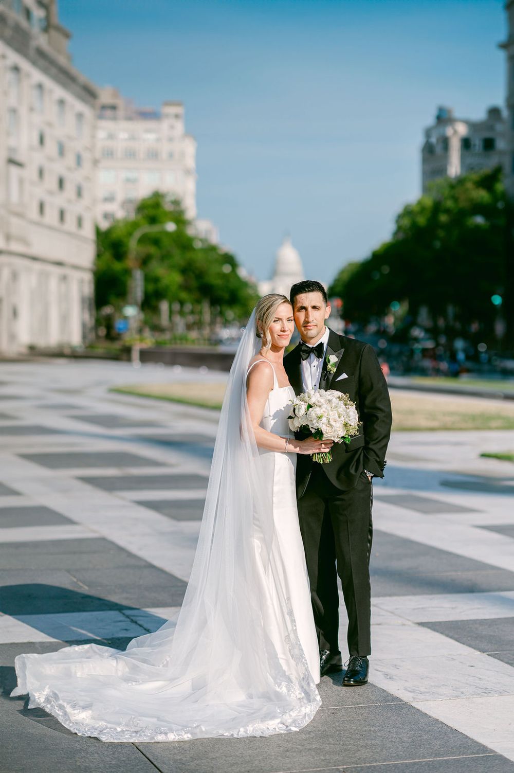 A romantic wedding couple poses in formal attire on a Washington D.C. plaza with a long flowing white veil and classic architecture.
