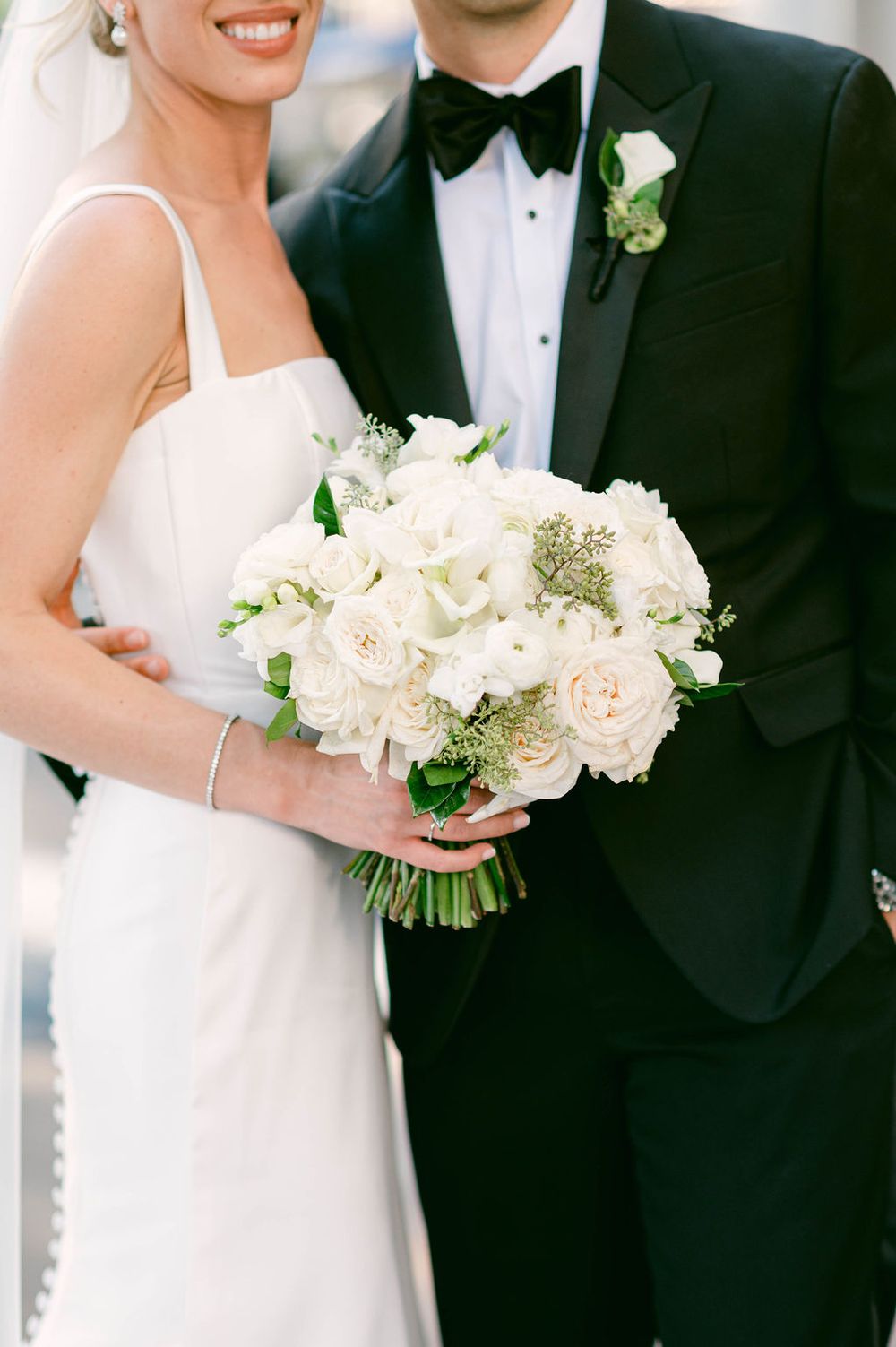 A bride in a white dress holds an elegant bouquet of white and blush flowers next to a groom in a black tuxedo.