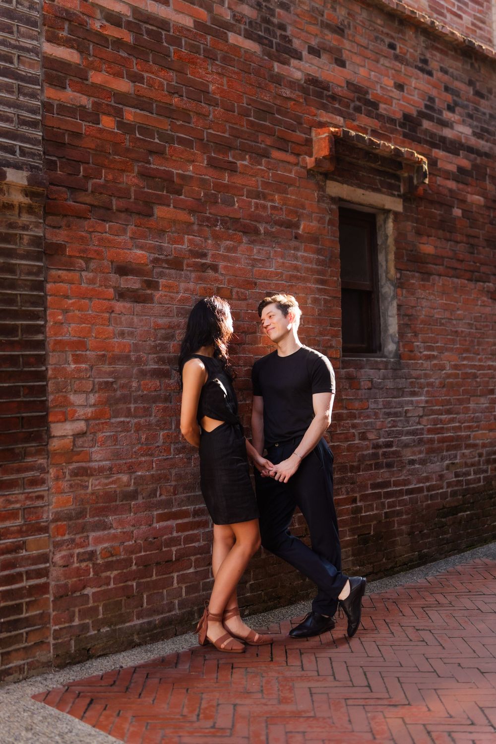 A romantic moment of a couple dressed in black against a rustic brick wall in an urban setting.