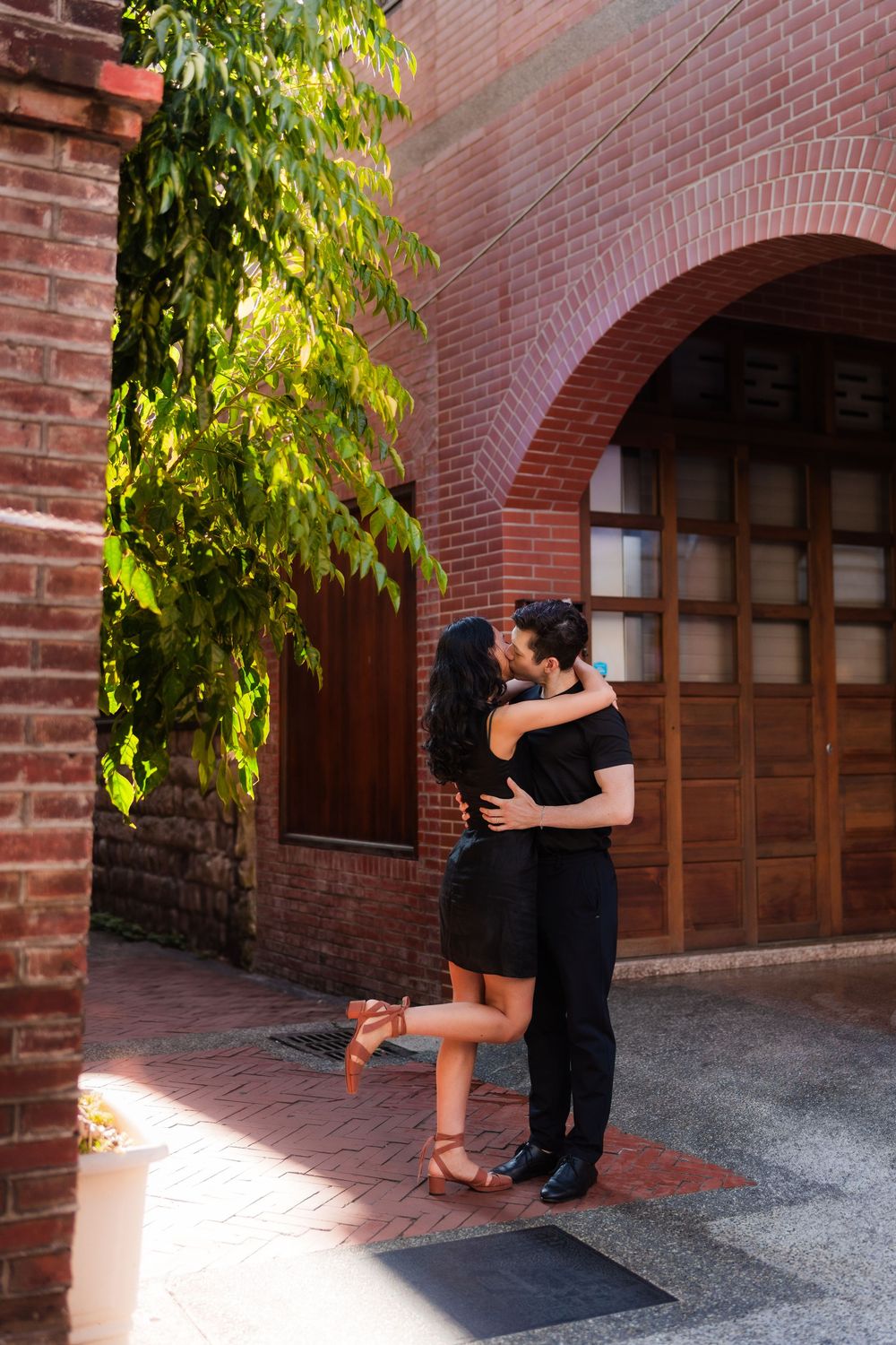 A couple shares an intimate moment against a red brick wall with arched doorways and green foliage.