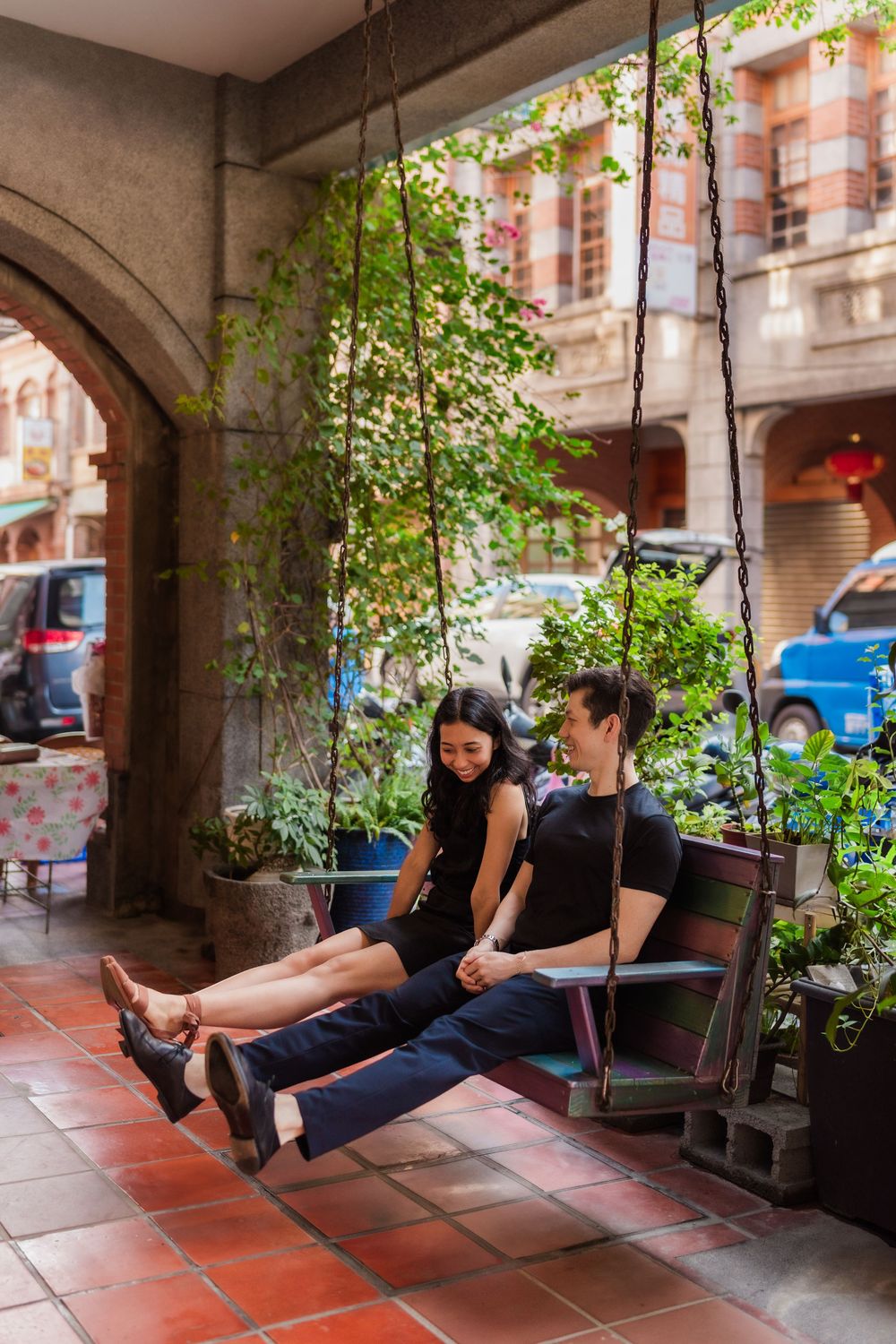 A cute couple in coordinating black outfits sit on a porch swing in a historic brick covered archway in Taiwan.