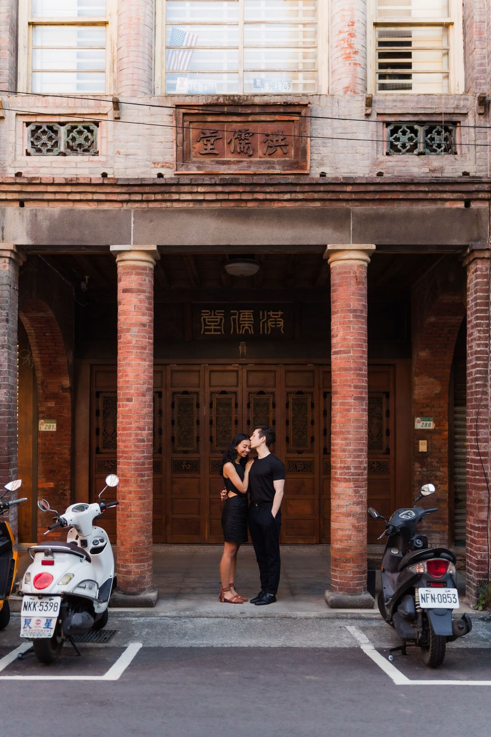 A couple standing in between historic looking brick columns near parked scooters and motorcycles lined up.