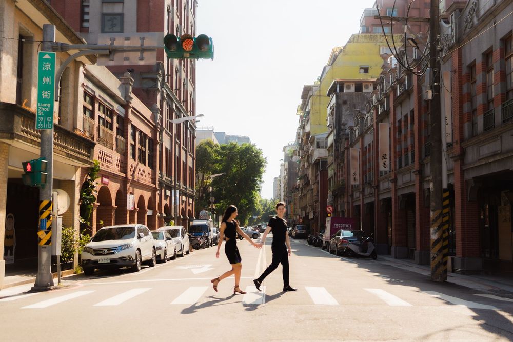 A couple crosses a sunny street in a historic urban neighborhood with brick buildings and parked cars  near Taipei's Dihua Street.