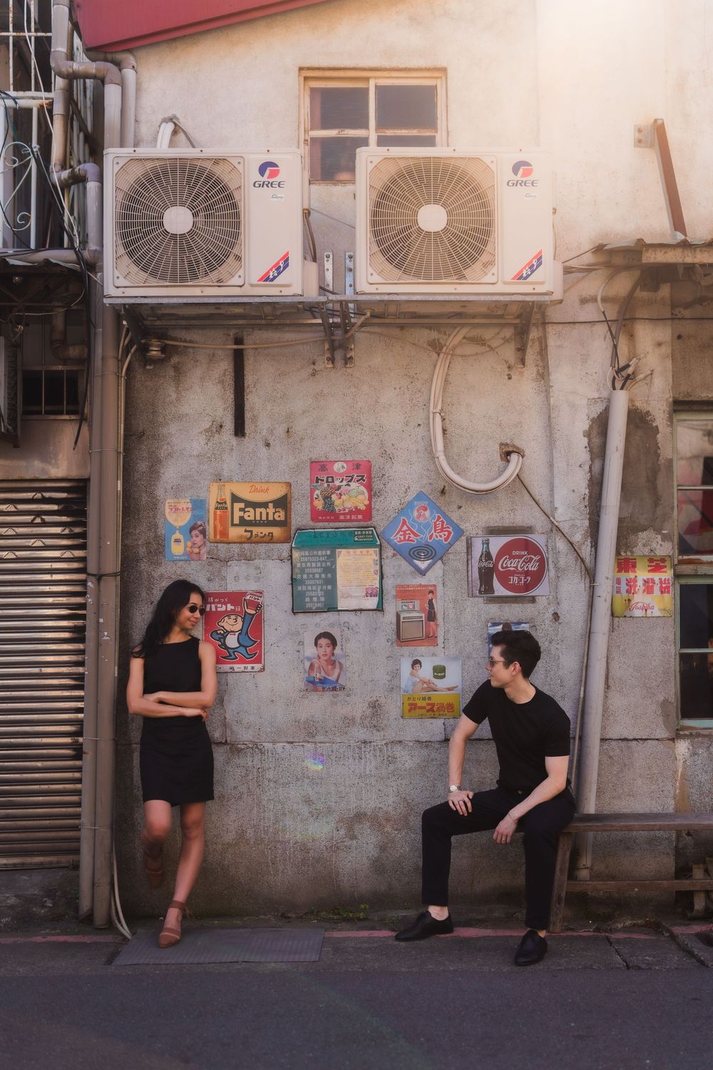 Two people in black clothing sit and lean against a grungy aesthetic wall covered in posters and air conditioning units.