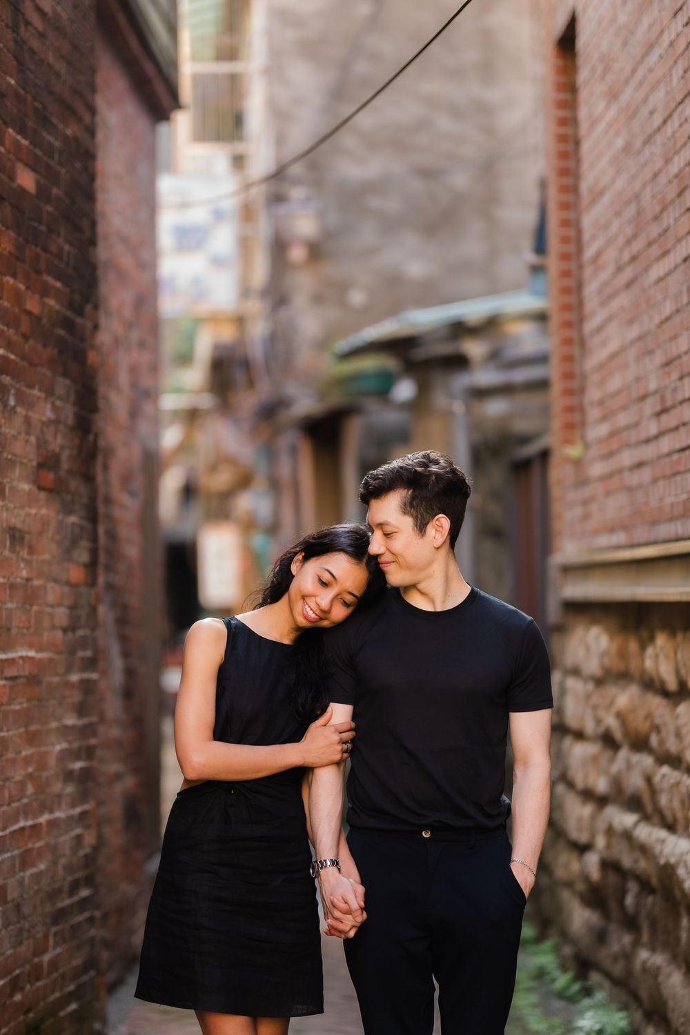 A couple in matching black outfits share a cute side embrace along a brick alleyway.