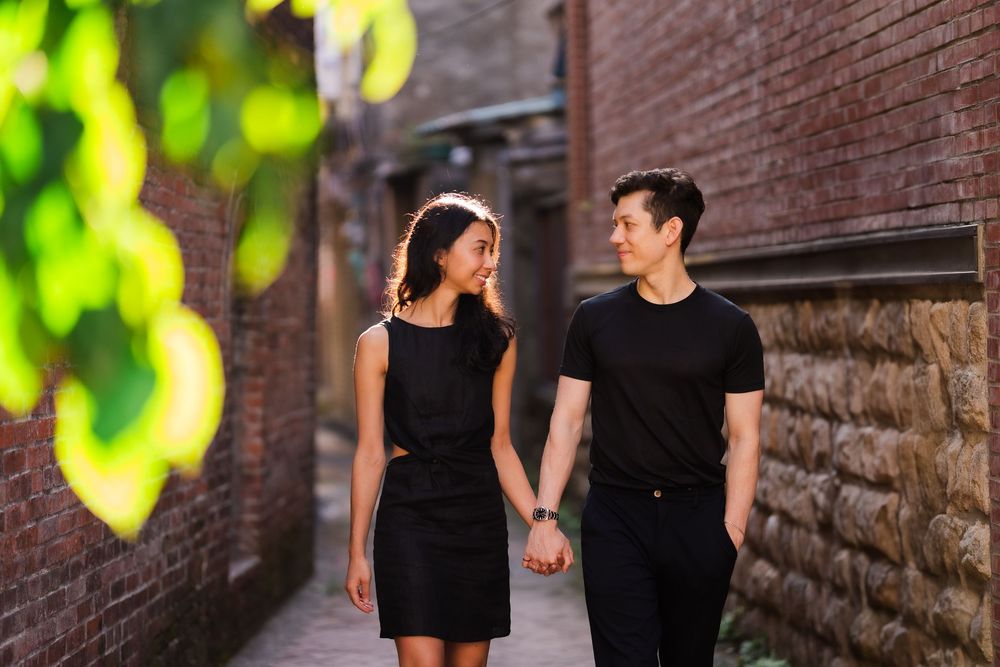 A couple in matching black outfits walks together along a sunlit brick pathway with green foliage.