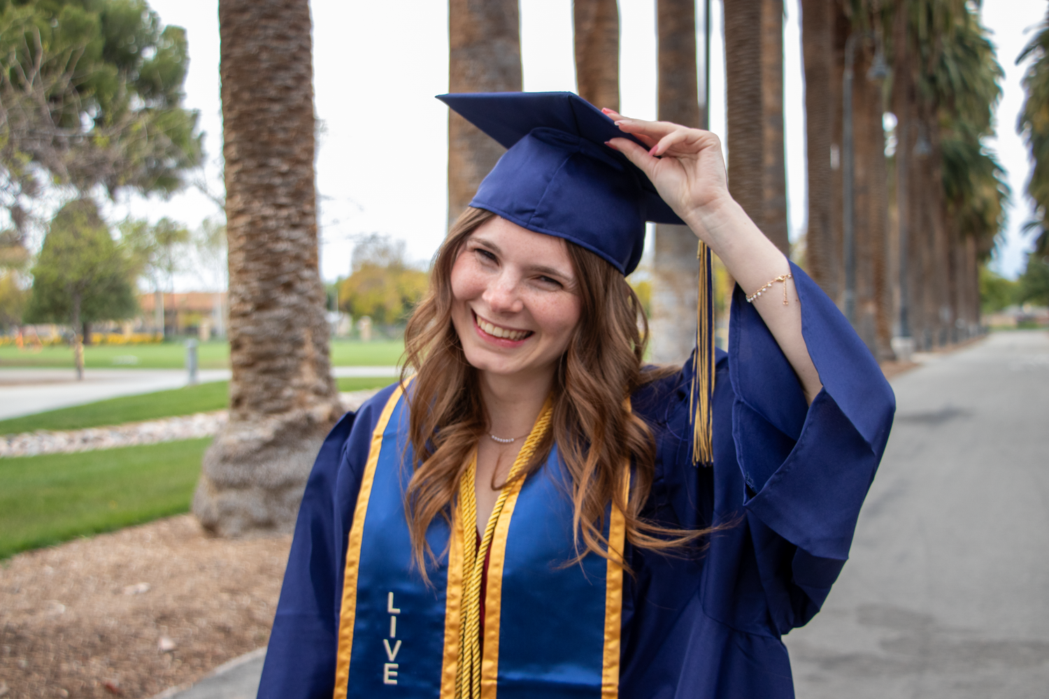 Smiling graduate in blue cap and gown holding her mortarboard outdoors among palm trees.