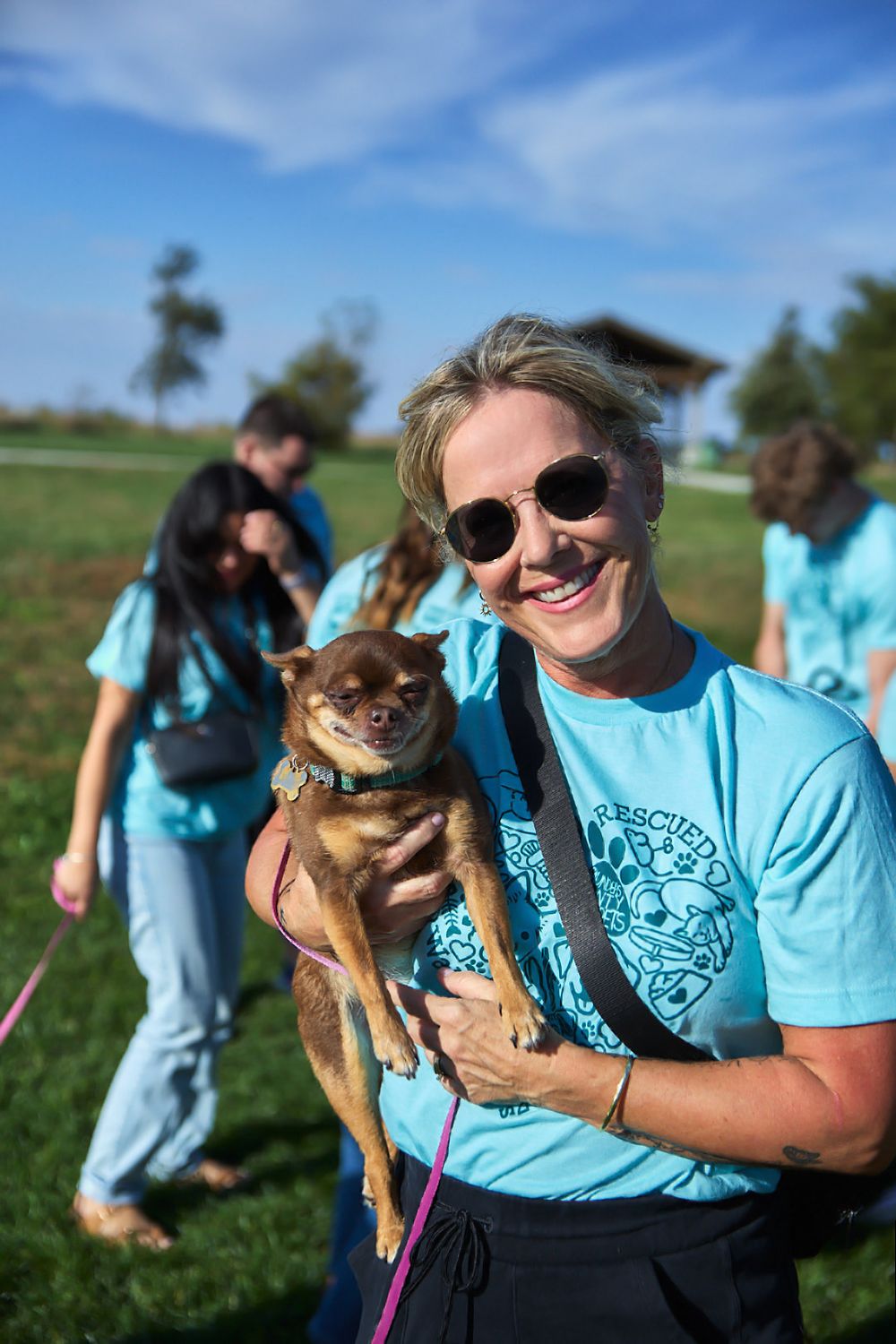 Person in light blue shirt holding a small brown dog at an outdoor event.