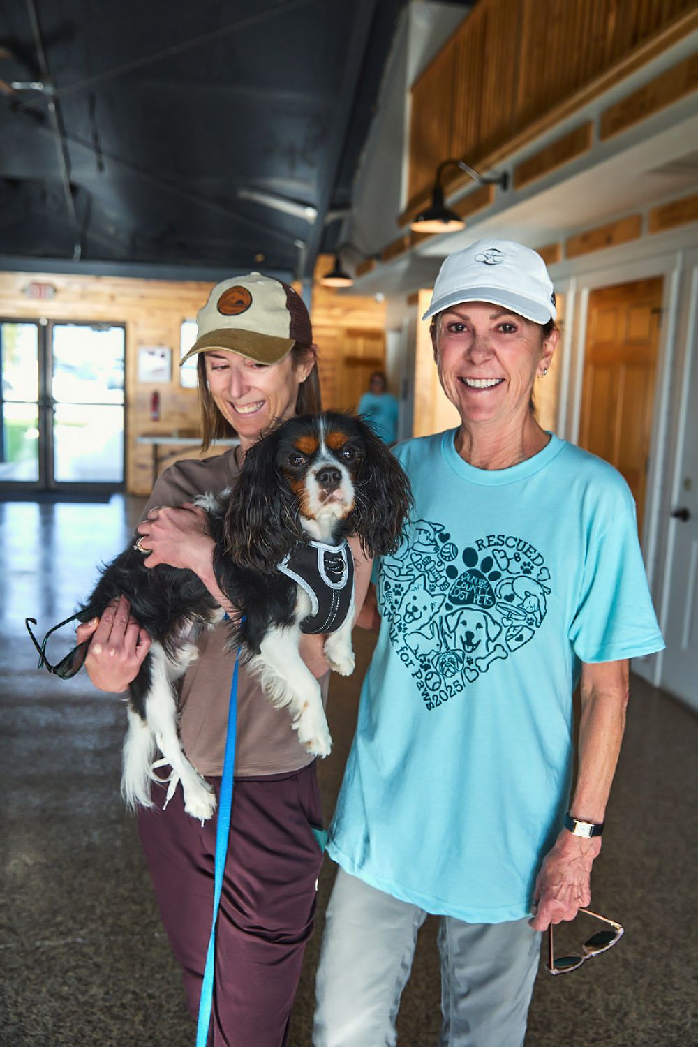 Small dog being held while people in turquoise shirts smile at an indoor gathering.