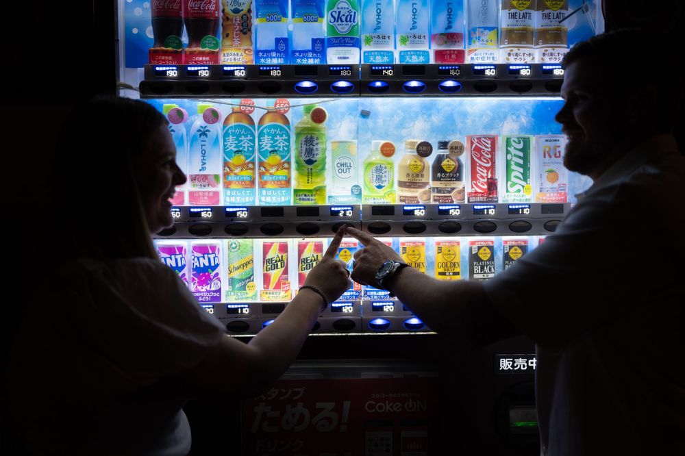 Couple silhouetted against a bright Japanese vending machine selecting drinks at night.
