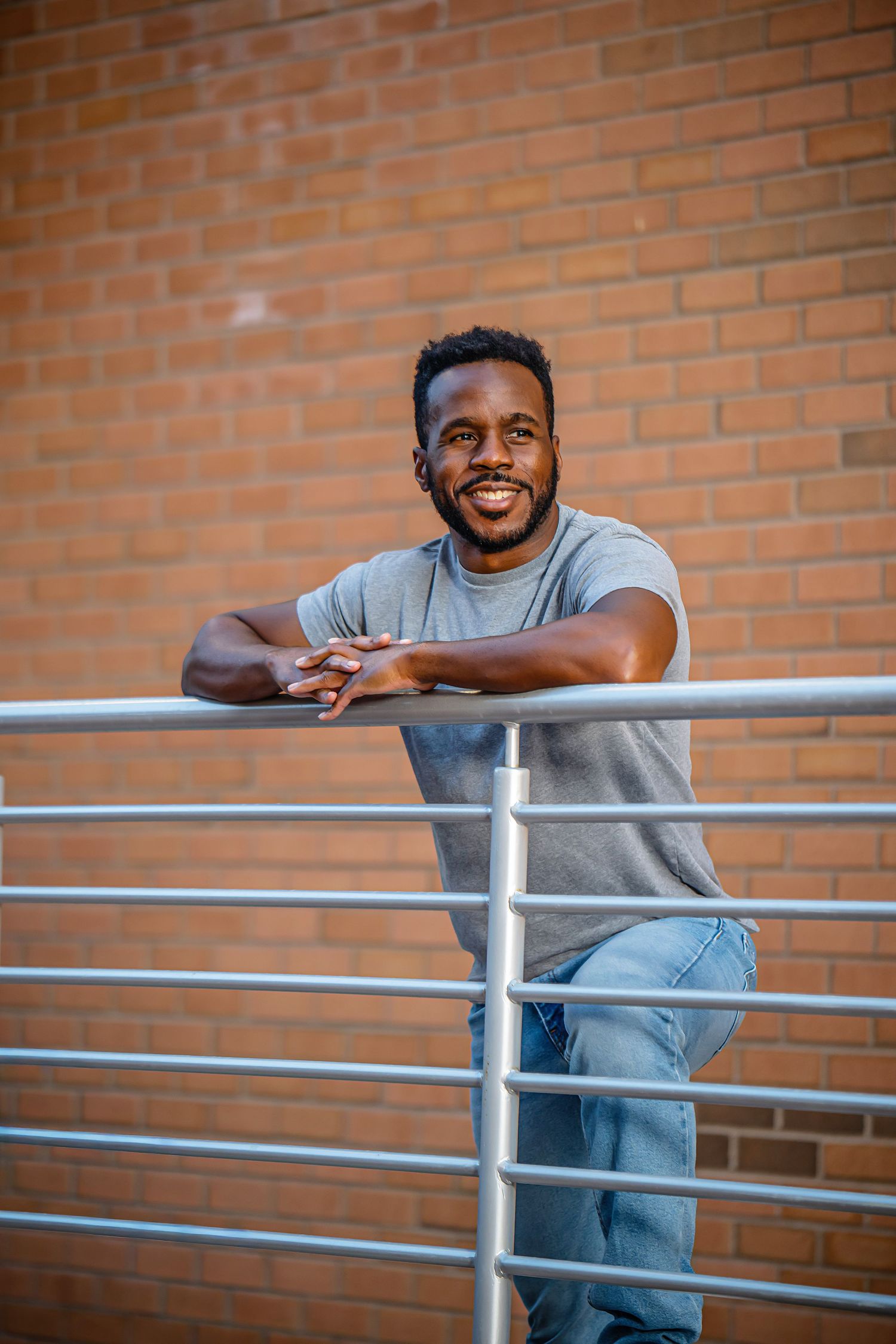 Person in casual grey t-shirt and jeans leans on metal railing against brick wall background.