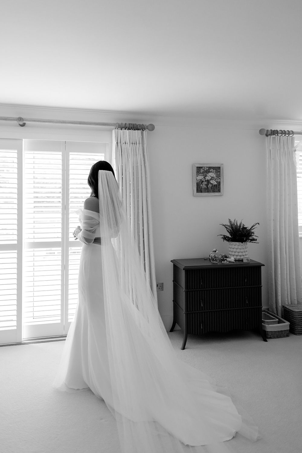 Black and white photo of a wedding dress with flowing veil in a bright room with white curtains and decor.