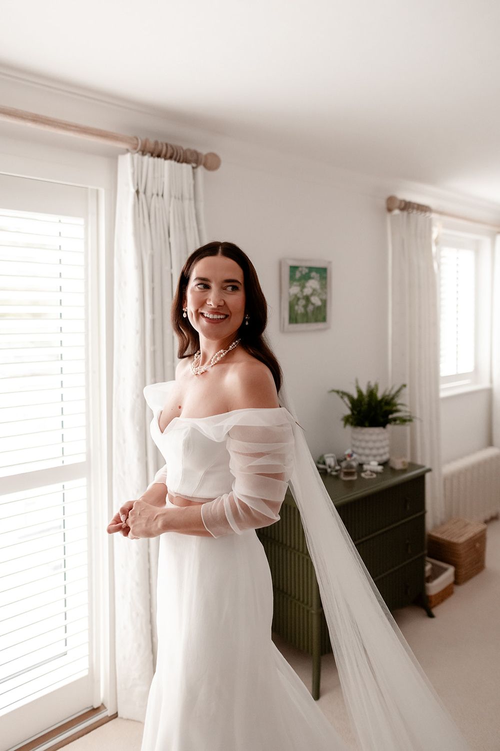 Elegant bride wearing an off-shoulder wedding gown with long veil in a bright white room.