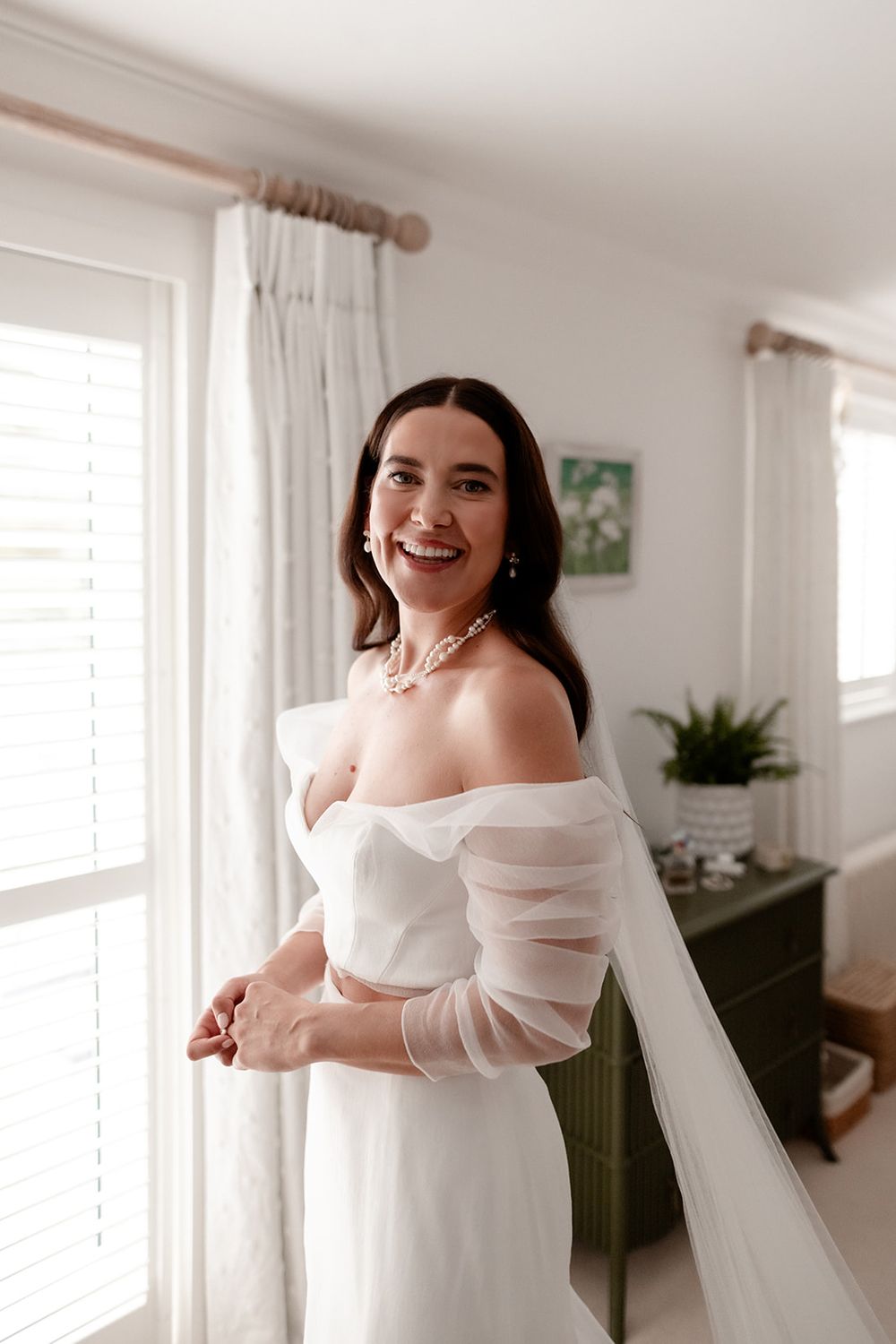 Joyful bride in an off-shoulder wedding dress with sheer sleeves poses by a window.