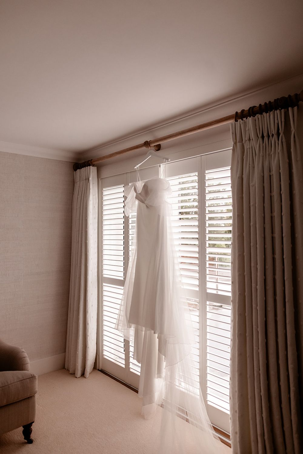 Wedding dress hanging by window with natural light streaming through shutters.