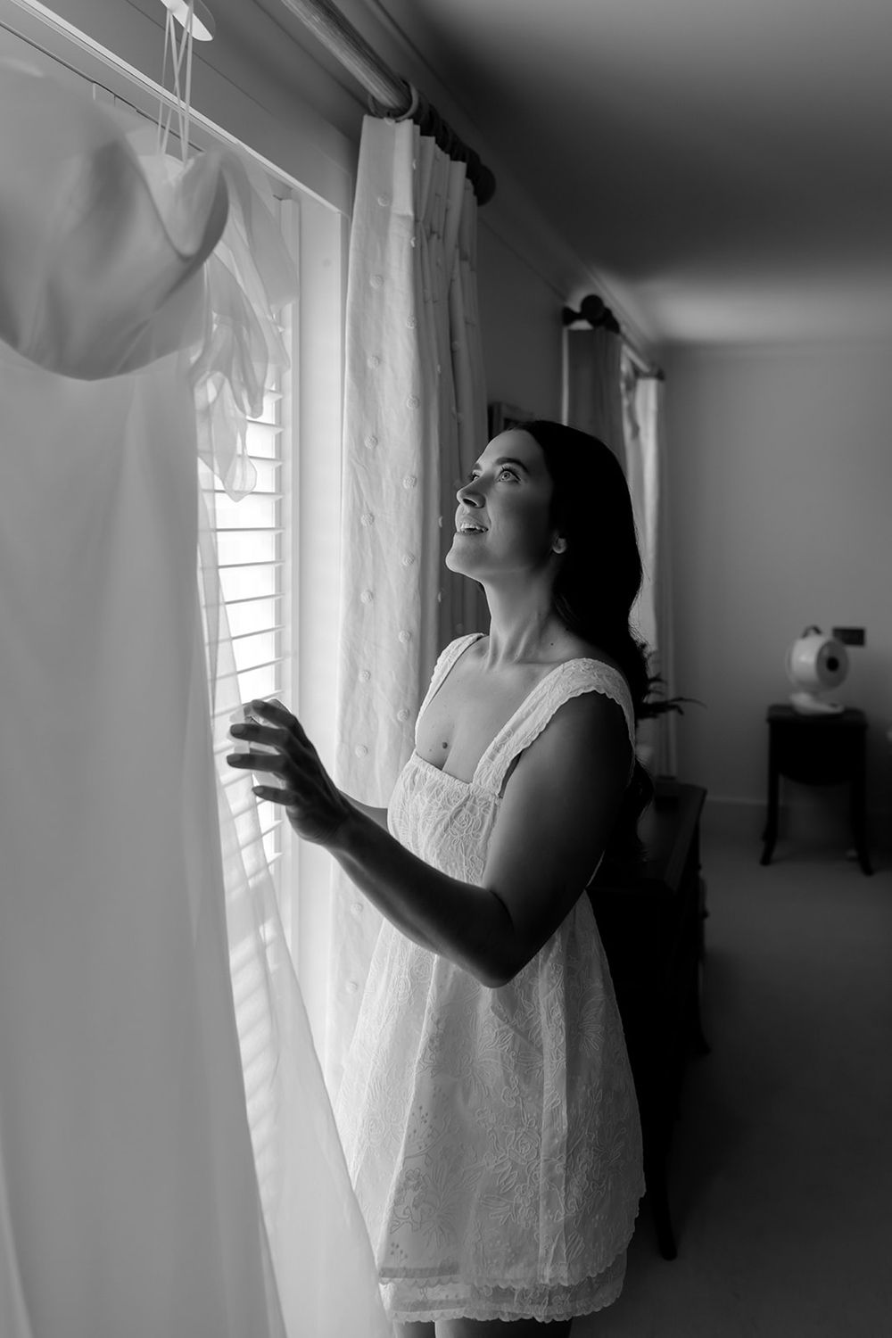 Figure in white lace standing by window curtains in dramatic black and white lighting.
