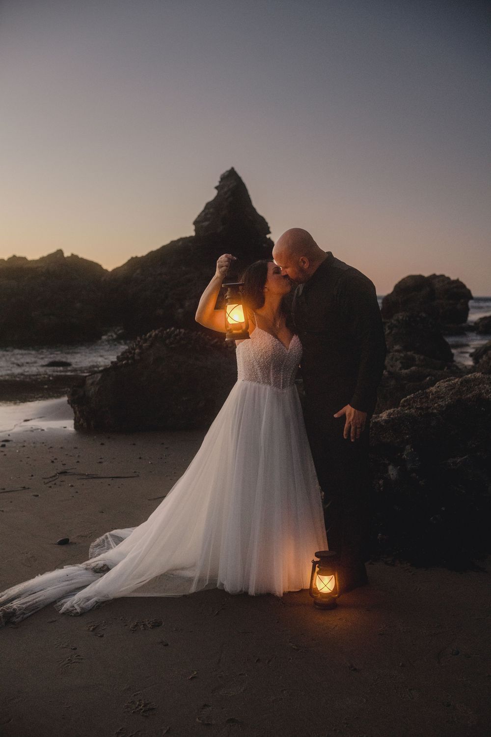 Romantic beach wedding photoshoot at sunset with lantern lighting and rocky coastal backdrop.