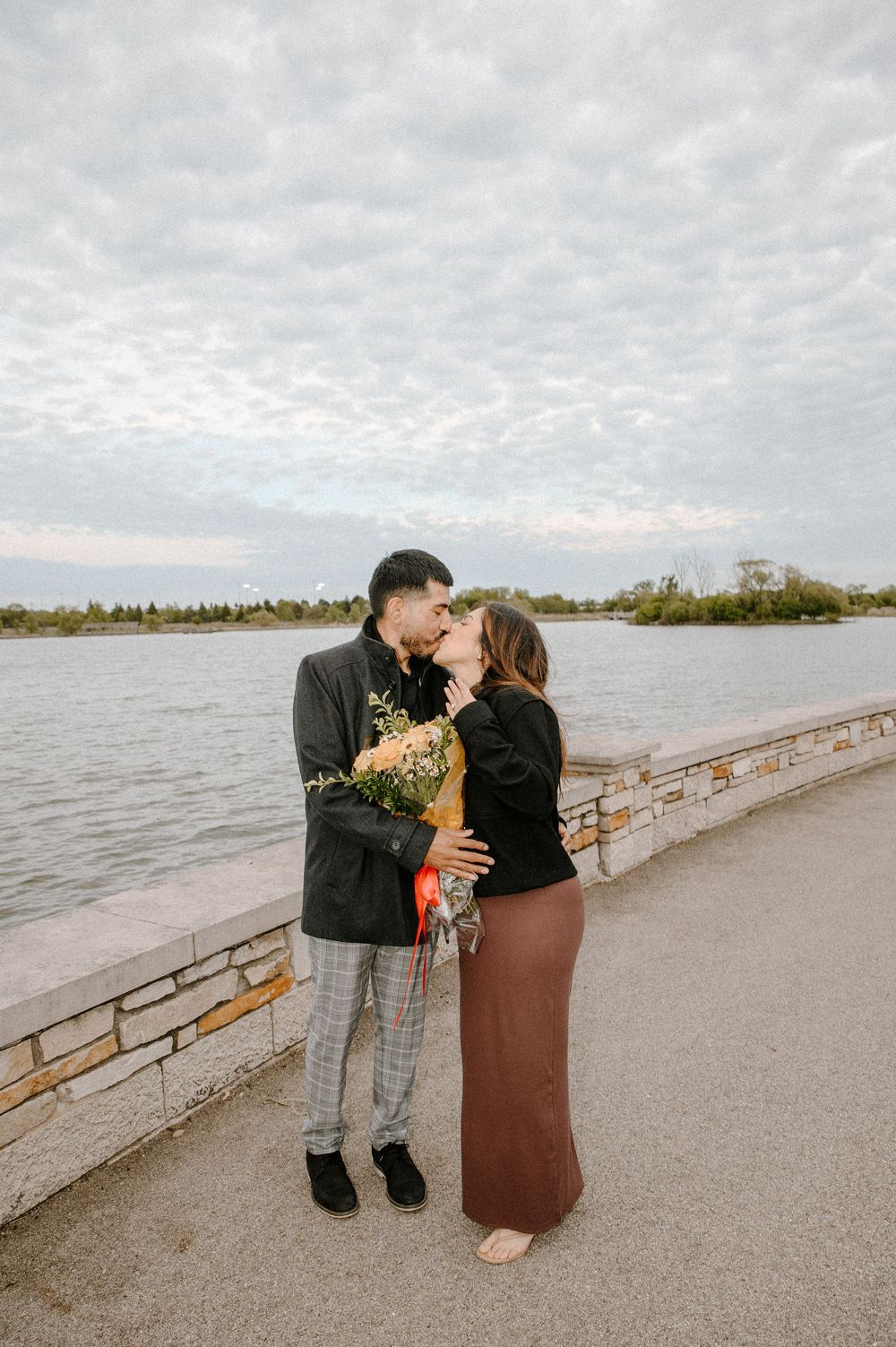 A romantic proposal moment unfolds on a cloudy day along a waterfront pier with a bouquet of flowers.