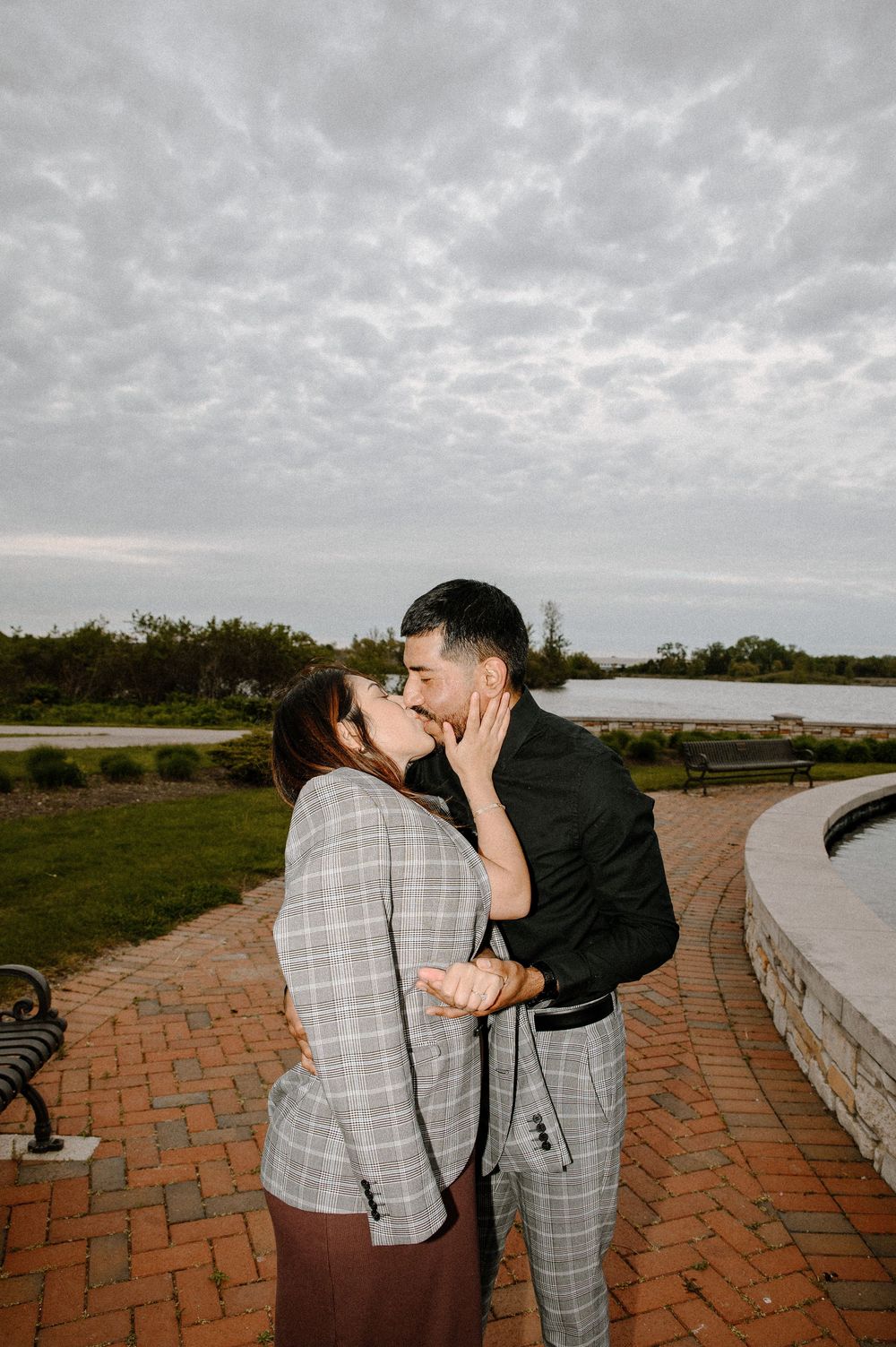 A romantic moment unfolds on a brick pathway by the water as a couple shares a tender kiss at dusk.