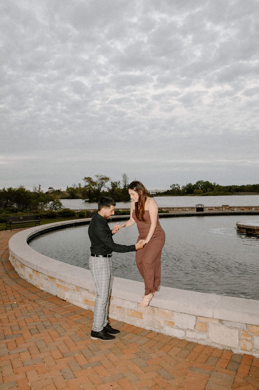 A couple shares an intimate moment on a curved brick walkway overlooking a peaceful waterfront on an overcast day.