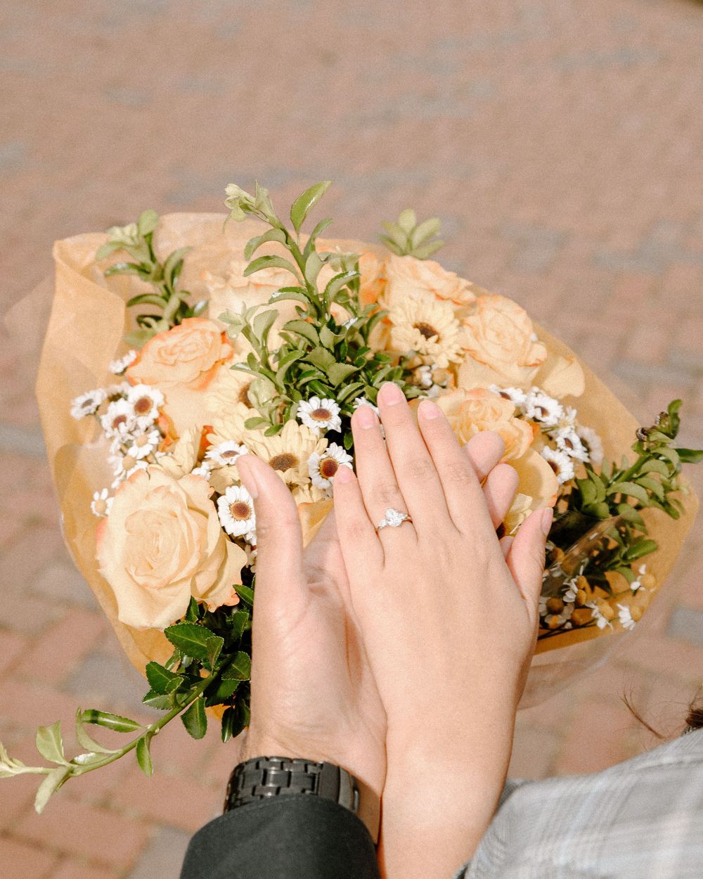 Two pairs of hands hold a delicate bouquet of peach roses and white flowers against a brick background.