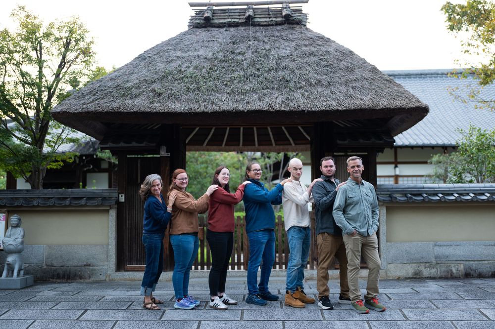 Seven people posing together in front of a traditional Japanese thatched-roof entrance gate.