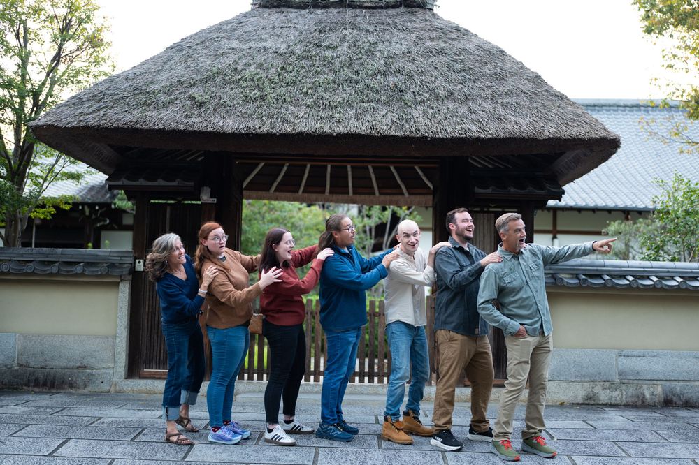 Group of seven people posing playfully in front of a traditional Japanese thatched-roof gate.