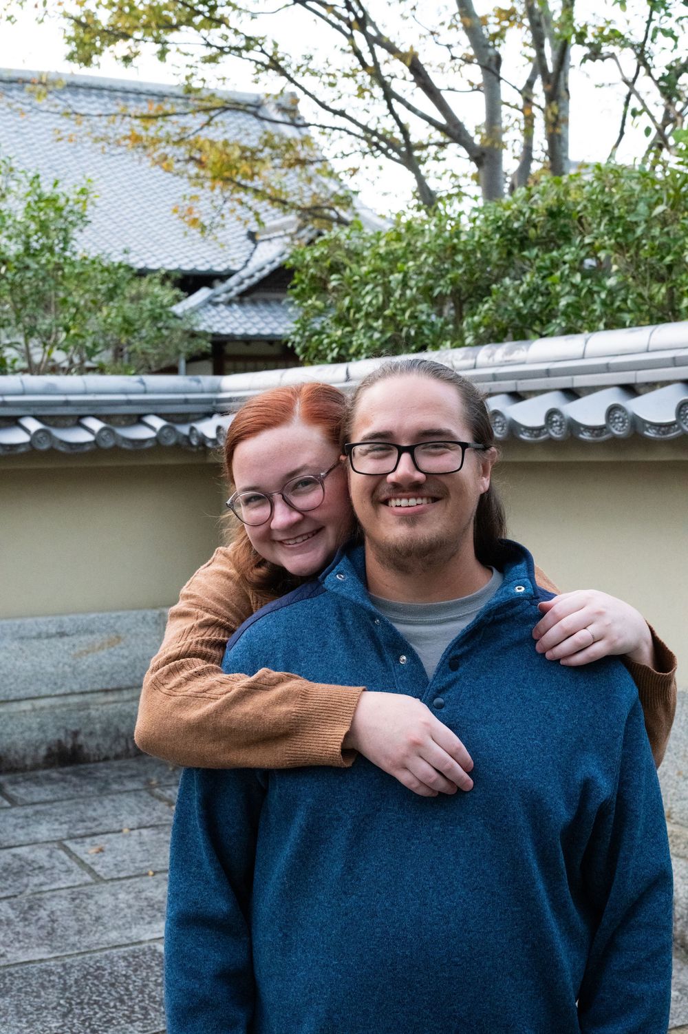 Smiling couple embracing outdoors near a traditional Japanese wall and rooftop scenery.