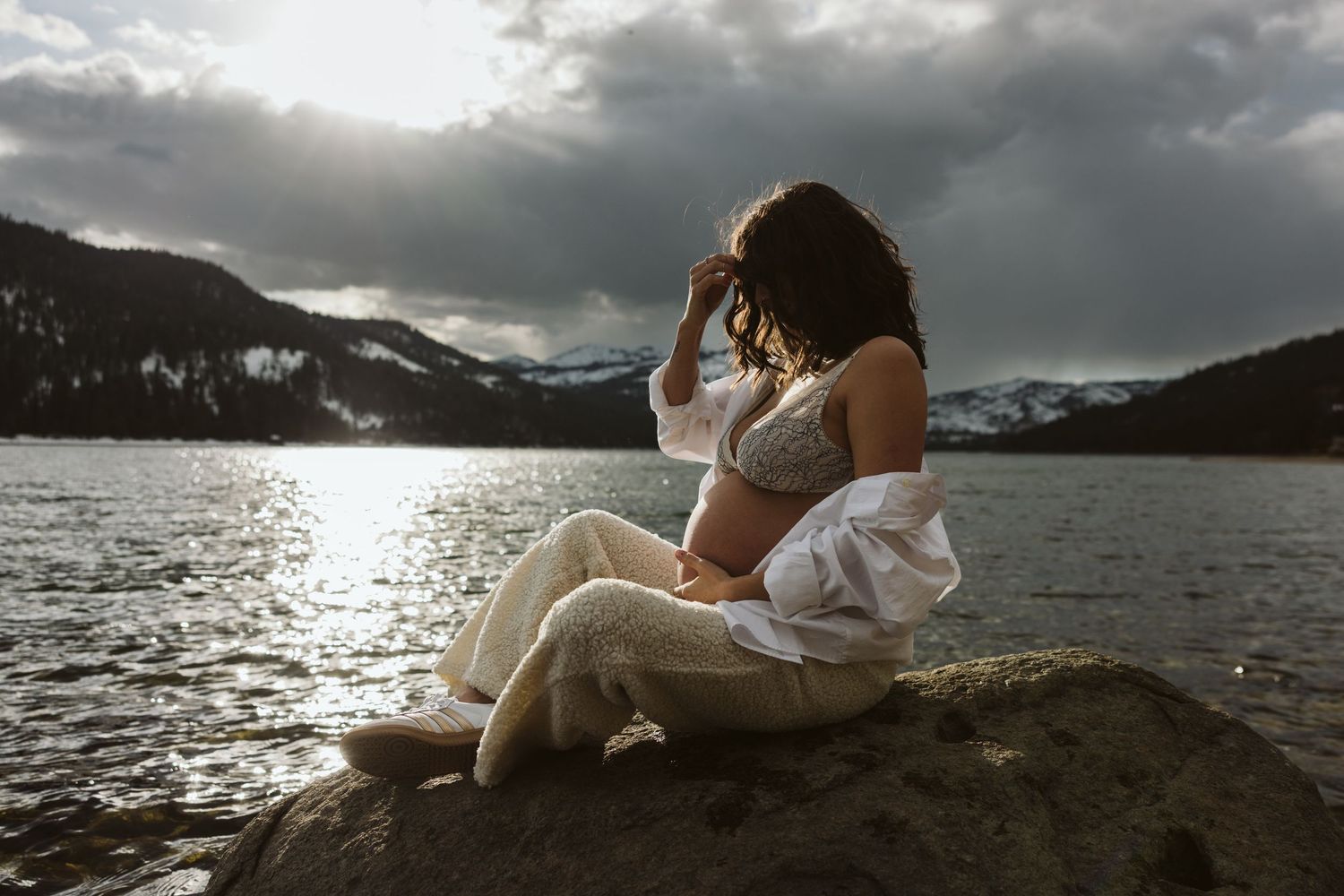 A couple in white outfits sits together on rocks by a lake at sunset with dramatic cloudy skies in the background.