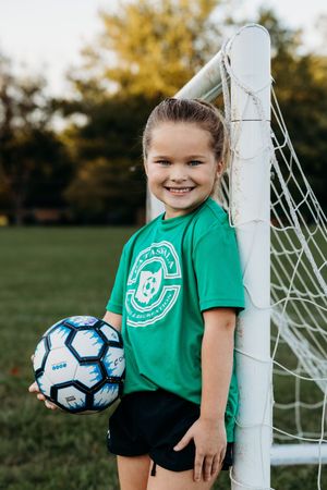 Young soccer player in green jersey stands by goal post holding blue and white soccer ball in outdoor field.