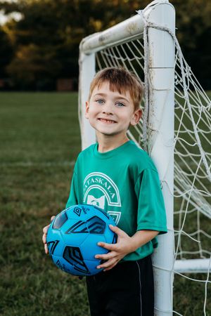Young soccer player in green t-shirt holds blue ball while posing by goal post on grassy field.