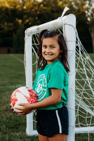 Young soccer player in green shirt and black shorts poses with ball by white goal post in outdoor field.