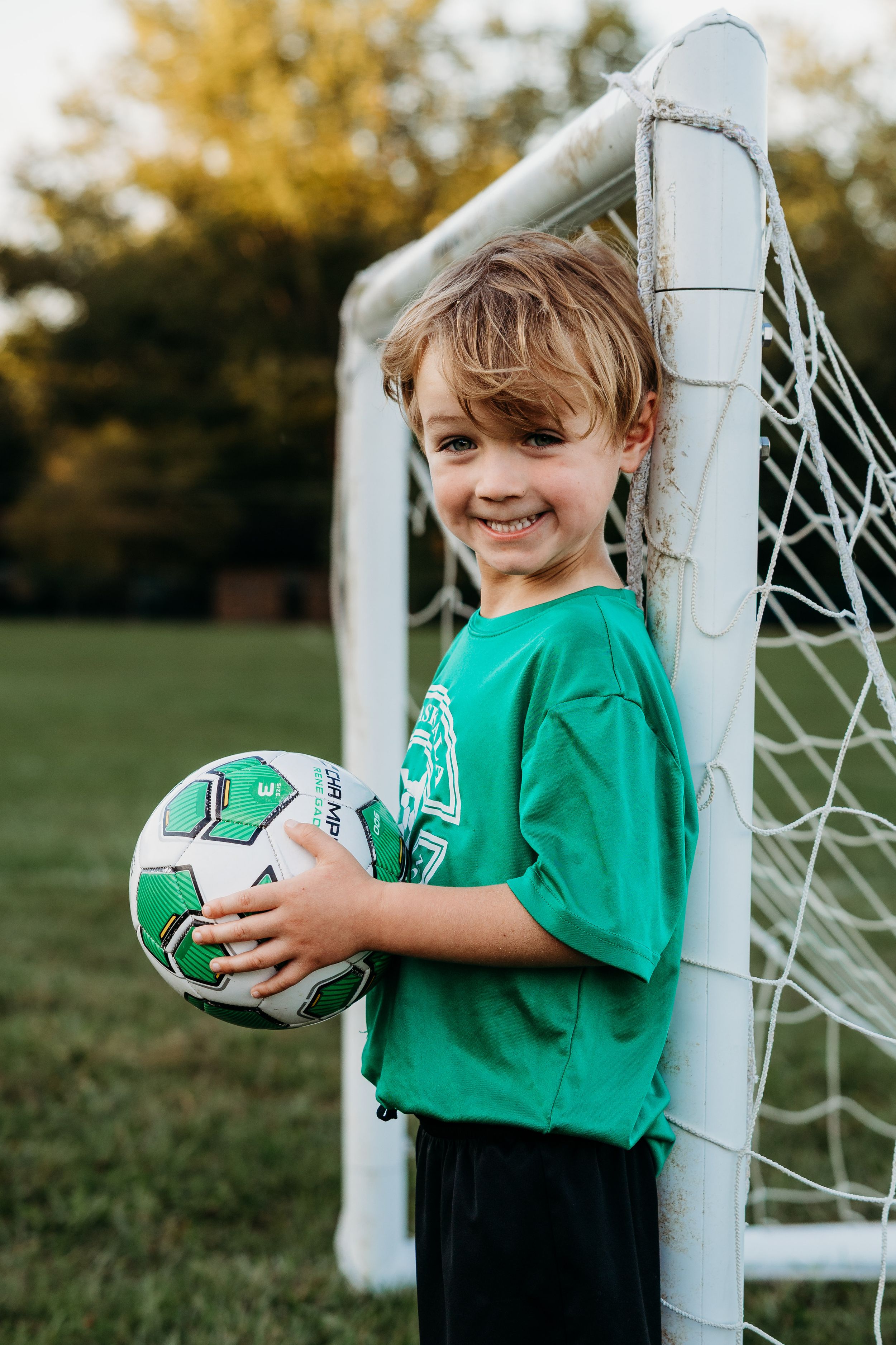 A young soccer player in a green jersey poses playfully with a soccer ball next to a white goal post.