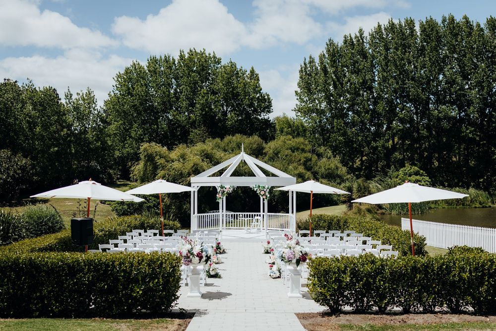 Outdoor wedding ceremony setup with white chairs and gazebo surrounded by lush green trees on a sunny day.