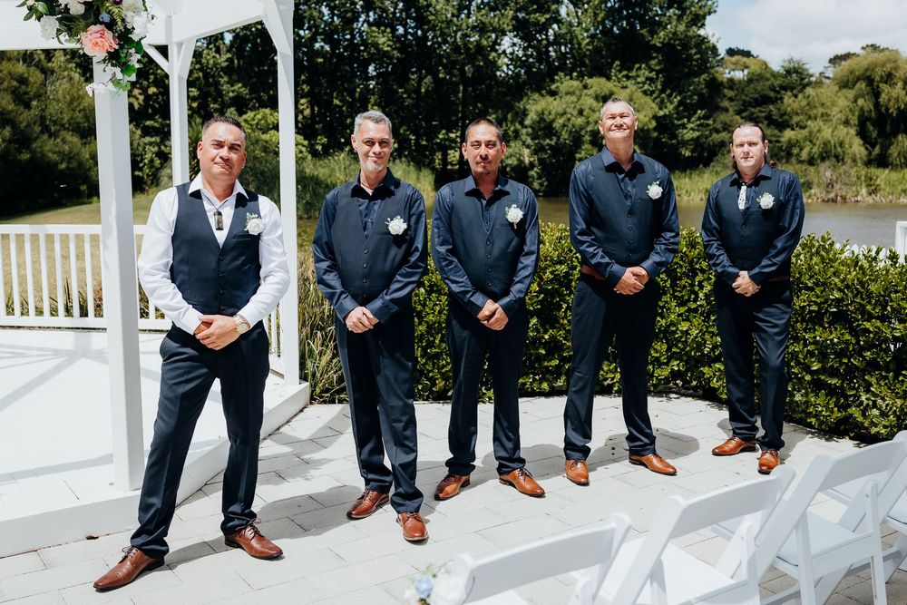 Wedding party members in navy suits and brown shoes lined up outdoors on white steps.