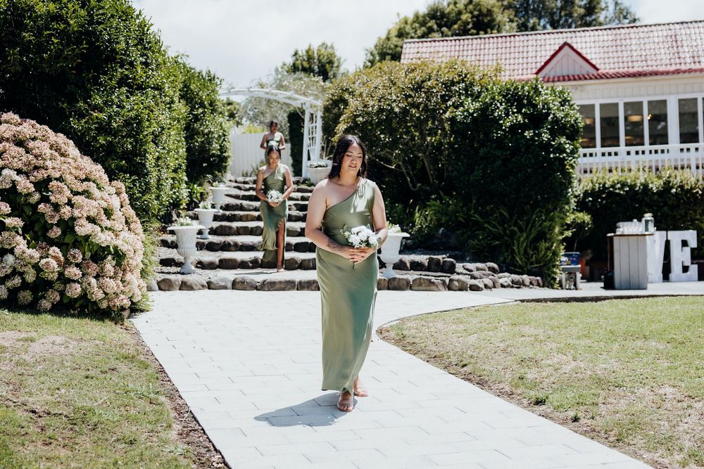 A bridesmaid in a sage green dress walks down an outdoor garden pathway with stone steps and hydrangeas.
