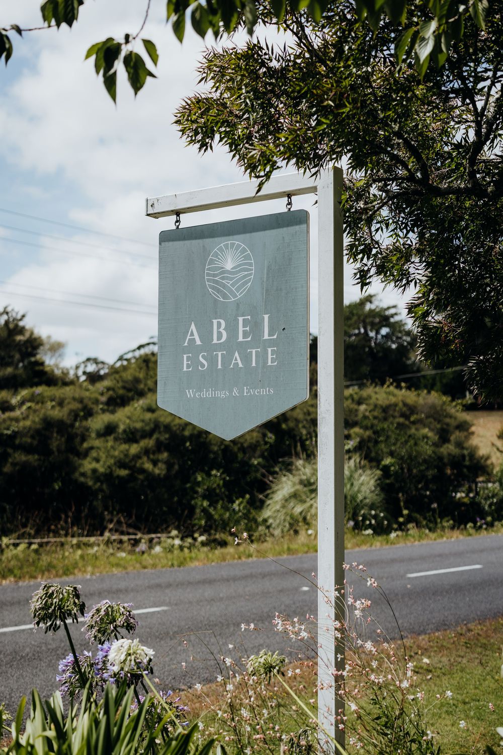 A white Abel Estate winery sign post stands along a rural road with green foliage and wildflowers in the foreground.