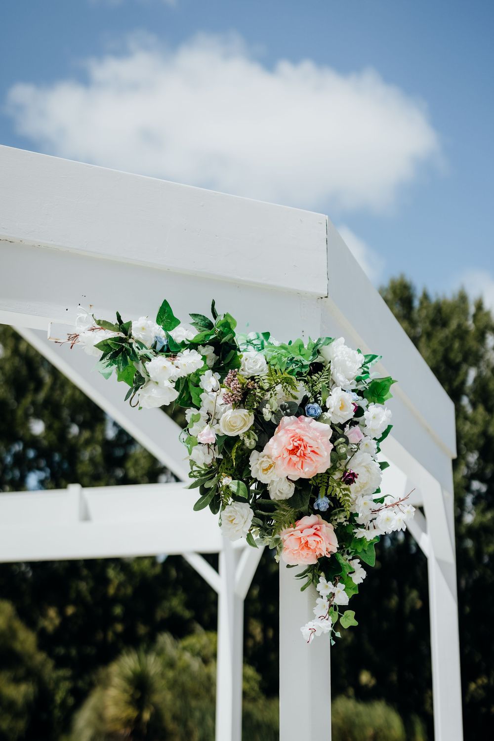 White wedding arbor decorated with delicate pink and white floral arrangements against a blue sky with puffy clouds.