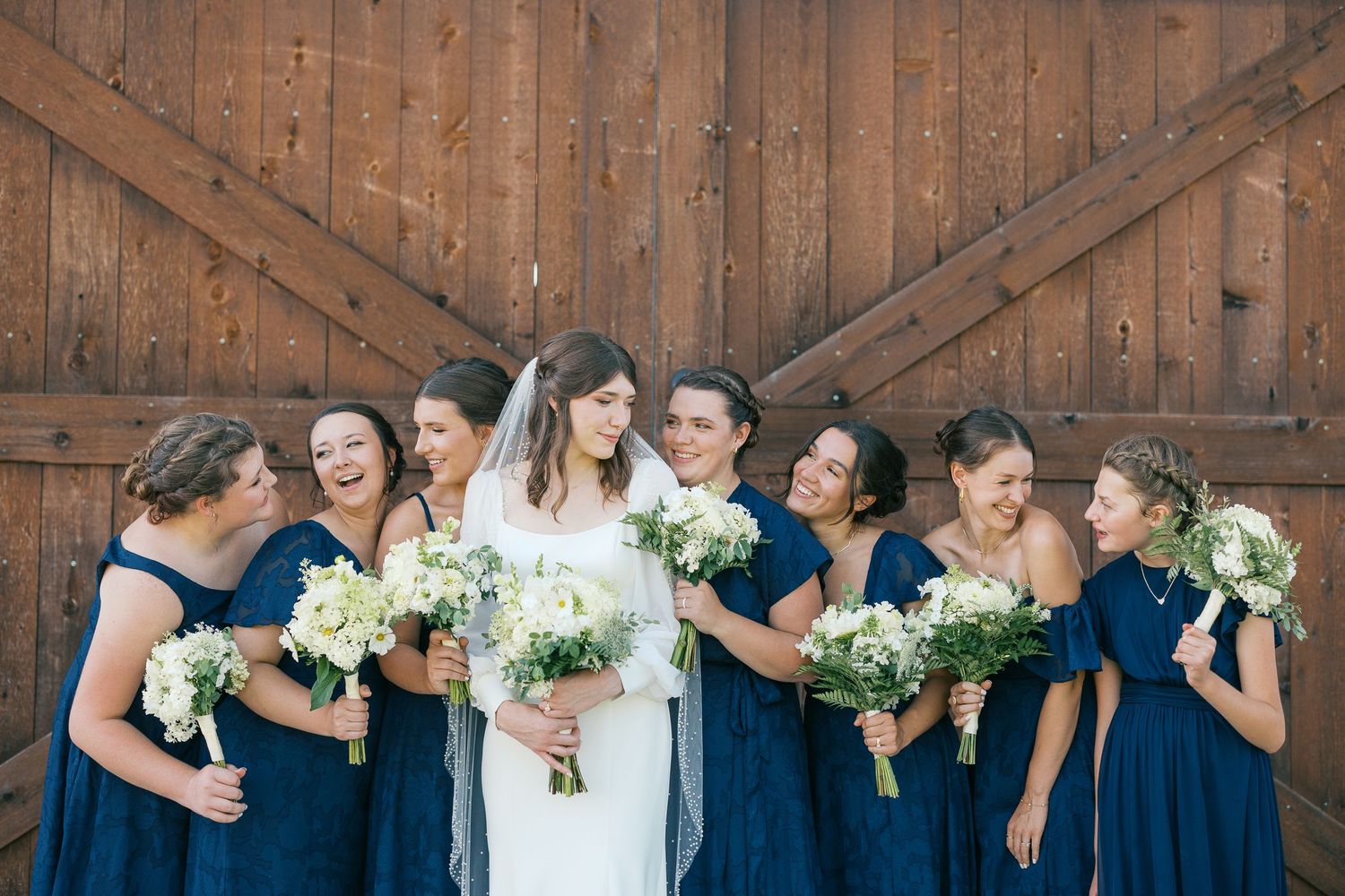 Wedding party in navy blue dresses poses together with white bouquets against rustic wooden barn door.