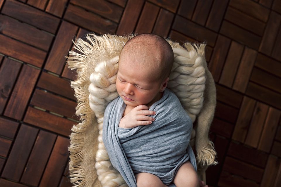 Artistic newborn photography with baby in soft gray wrapping against dark wood background.