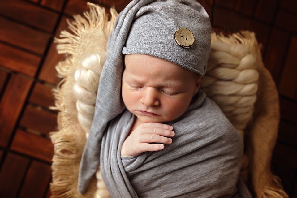 Newborn portrait with gray swaddle and matching cap on wooden surface with cream fur accent.