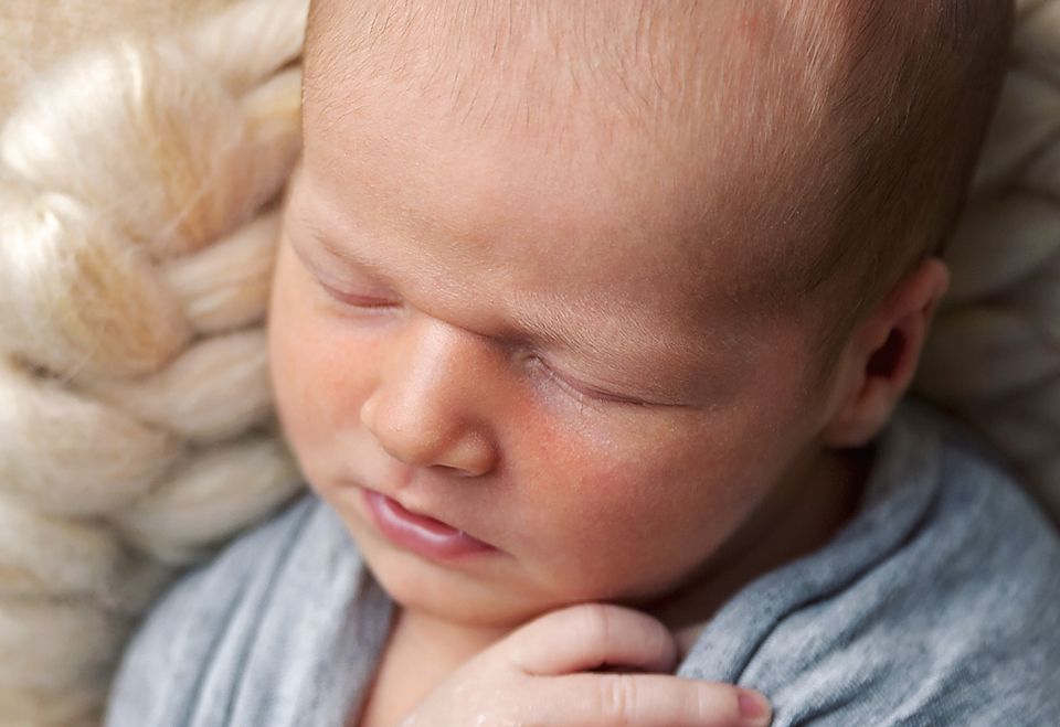 Gentle close-up portrait of sleeping baby in soft gray wrap against cream textured backdrop.