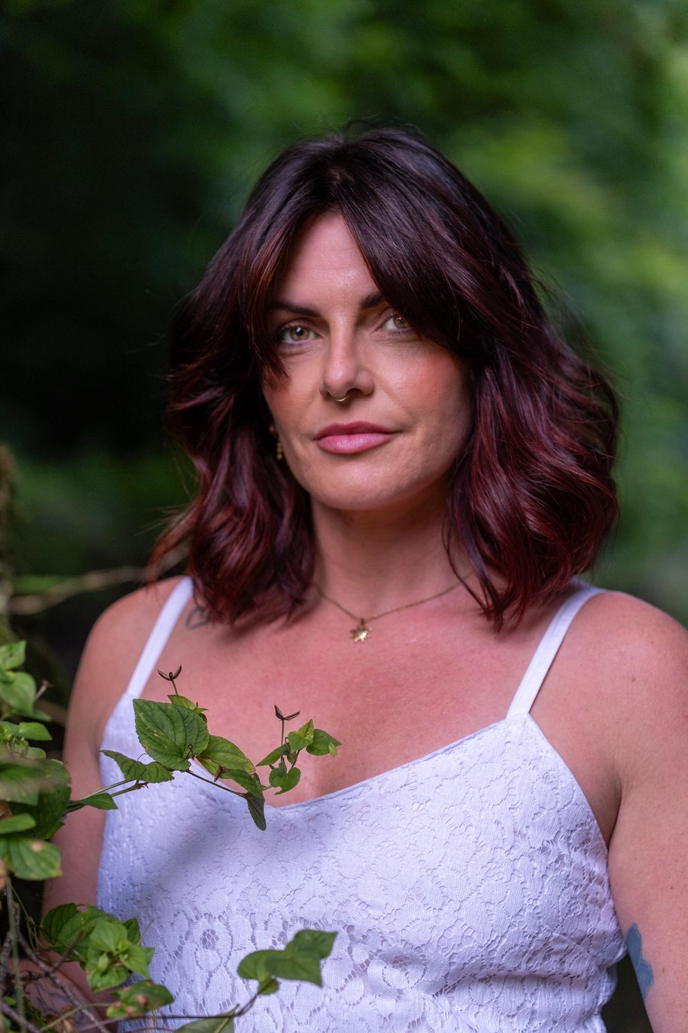 A person in a white lace camisole poses outdoors with dark wavy hair and green foliage in the background.