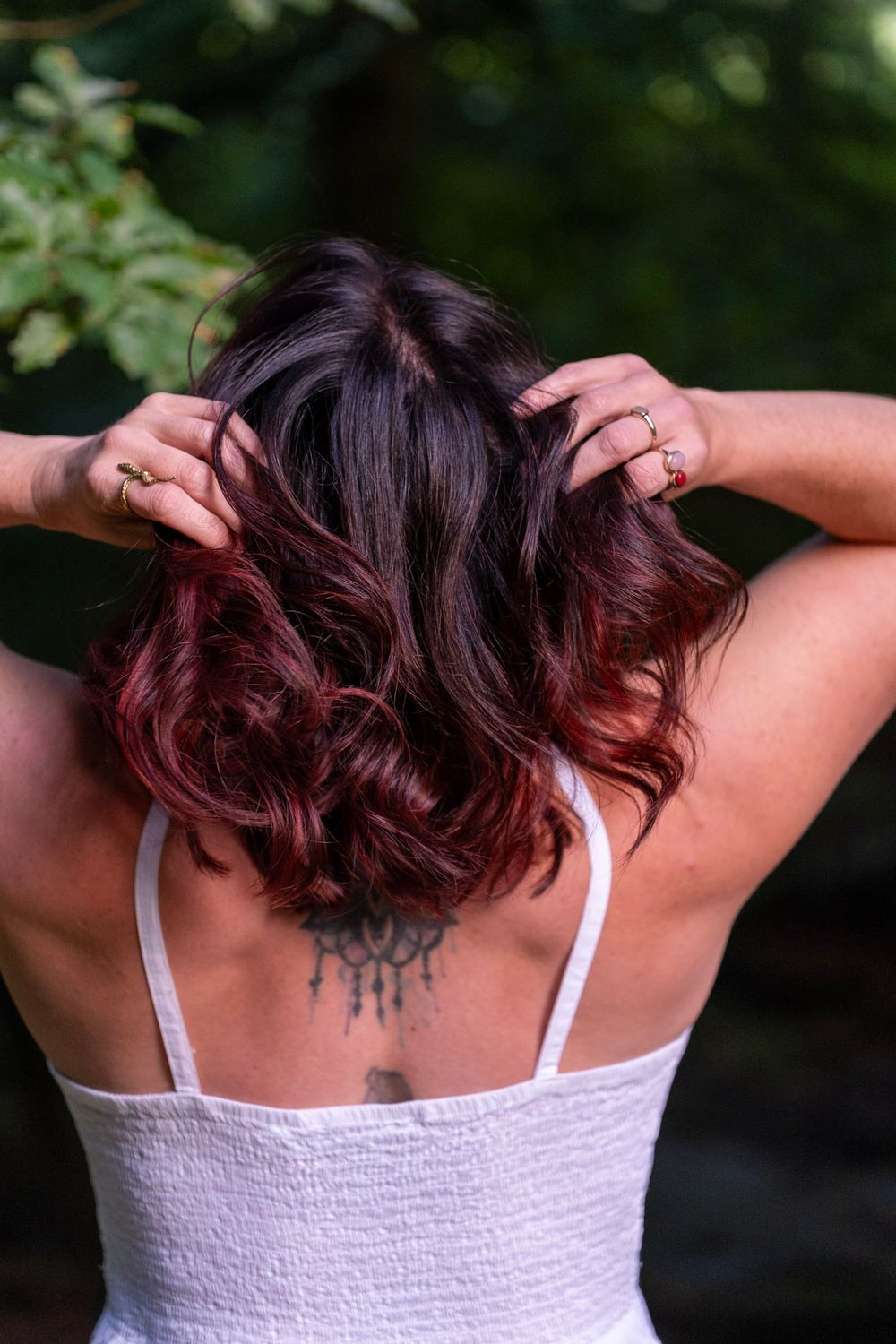 Back view of person with burgundy ombre wavy hair wearing white tank top showing small tattoo on upper back.