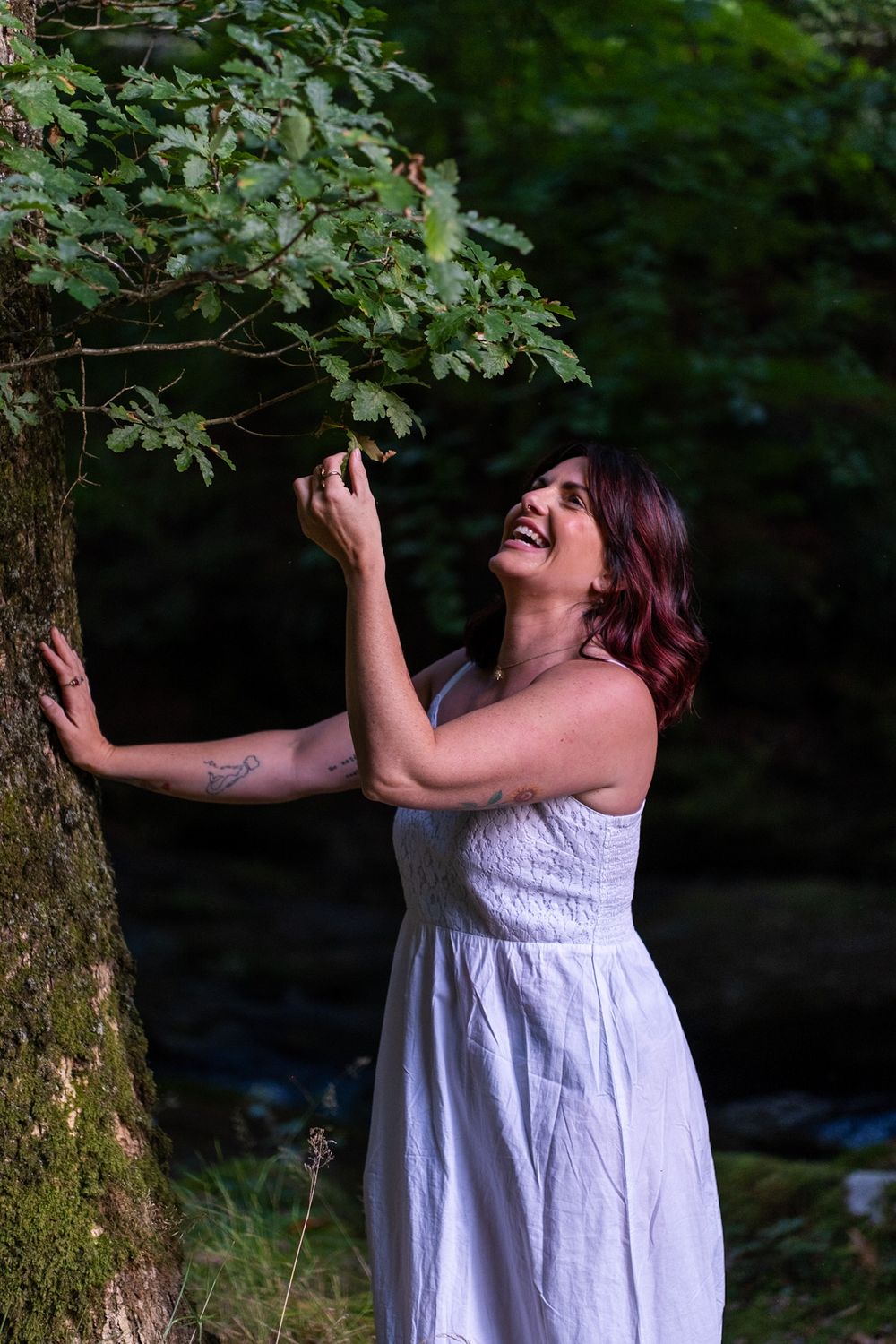 Someone in a flowing white dress laughs joyfully while reaching up to touch tree branches in a forest setting.