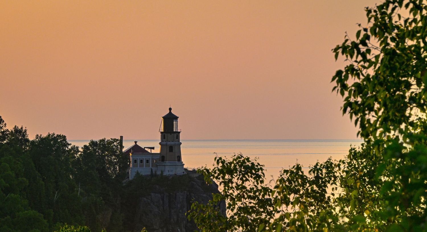 Panoramic sunset view of a tree-lined skyline with silhouetted lighthouse and golden sky in the background.