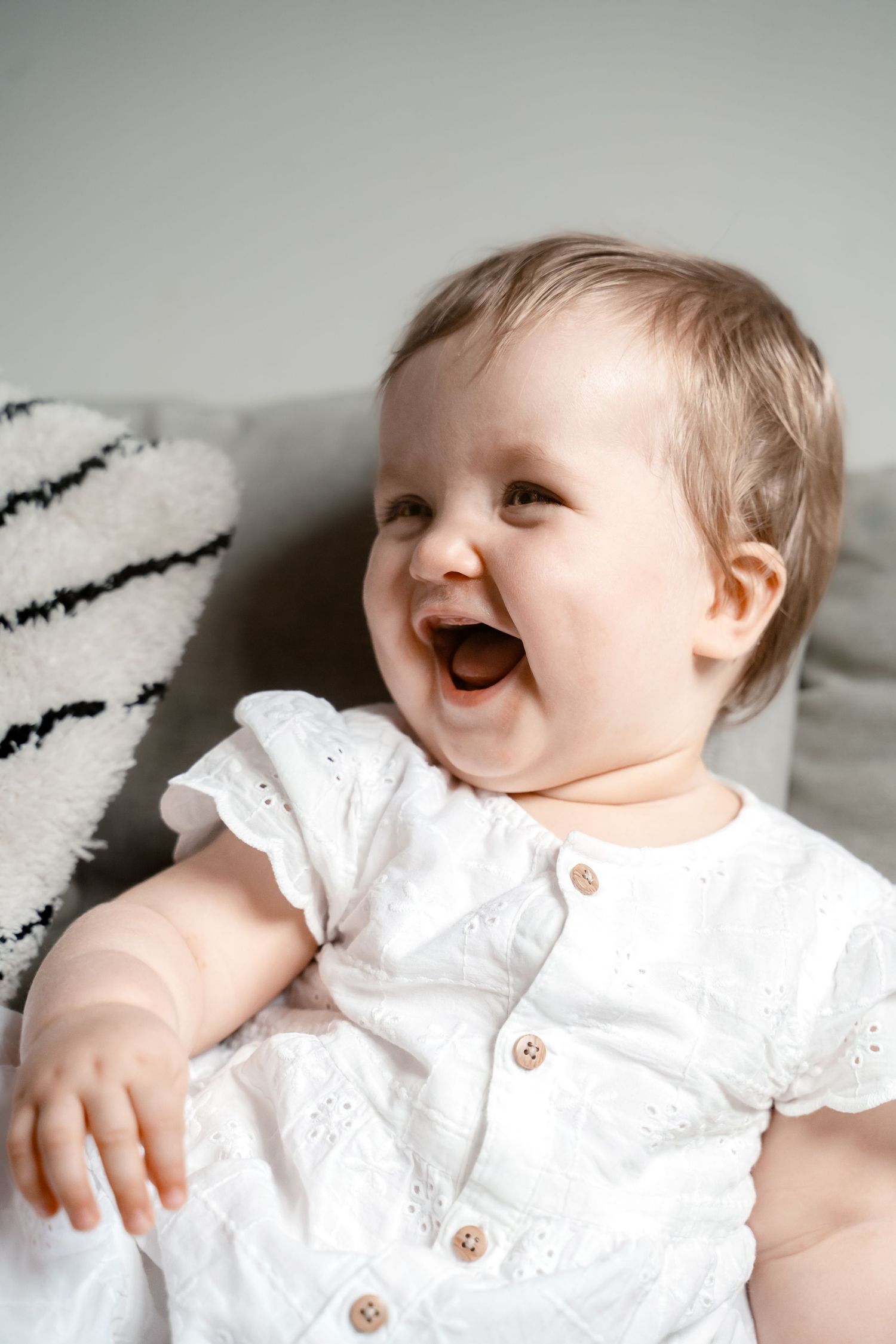 A baby wearing a white eyelet dress lies on white bedding while laughing joyfully in a series of connected photos.