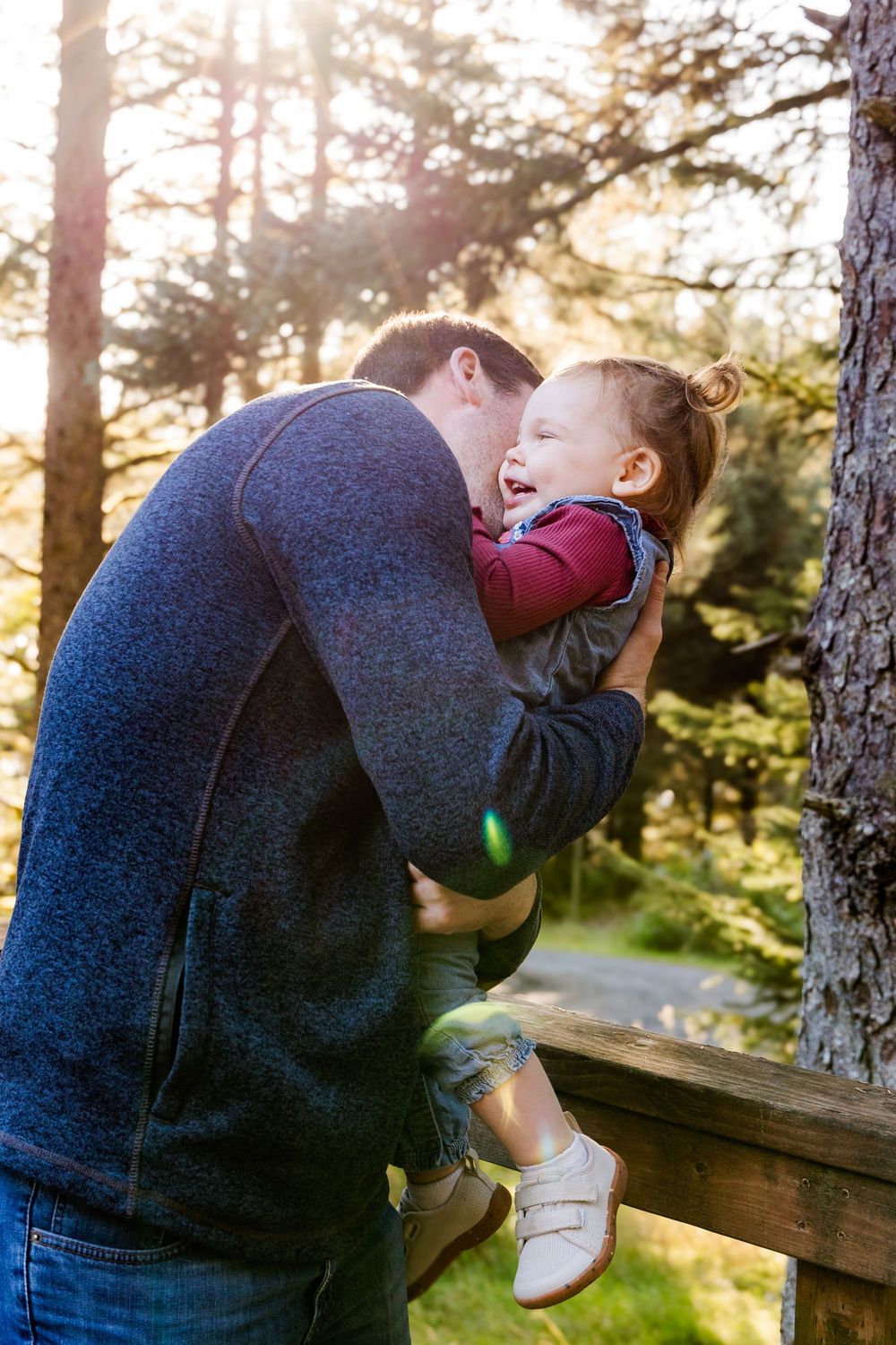 A parent leans over a wooden railing to hold a small child upside down playfully in a forest setting at sunset.