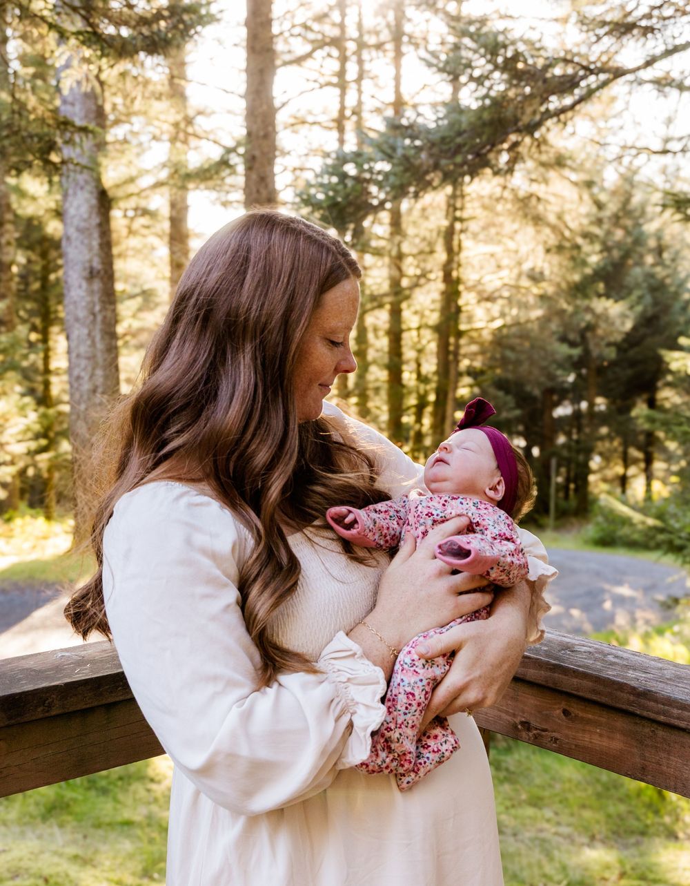 A parent in a white sweater lovingly cradles and kisses a baby in a floral outfit in a sunlit forest setting.